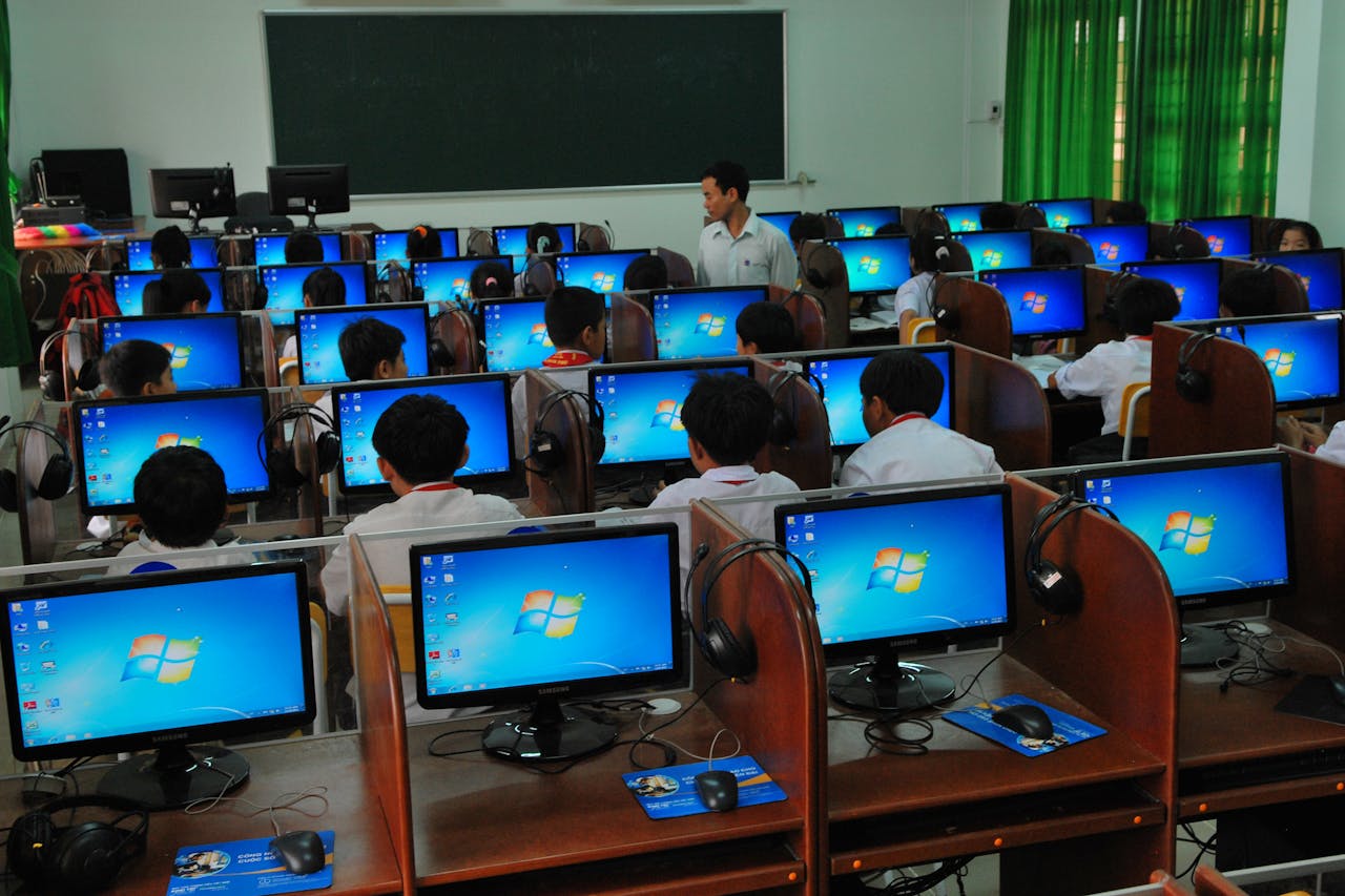 Students learning in a computer lab with multiple monitors and a teacher instructing.